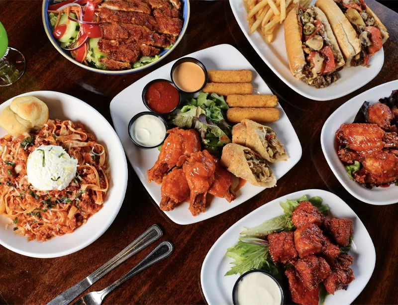 overhead view of plates of food including chicken wings, steak & cheese eggrolls, mozzarella sticks, bolognese pasta, steak & cheese sub with fries, and green salad topped with chicken
