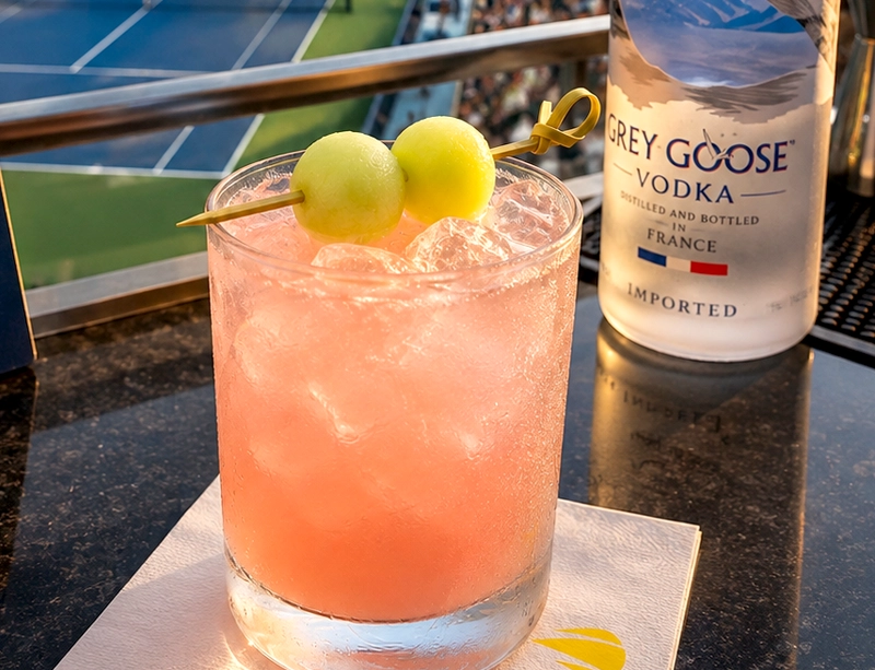 short pink cocktail over ice garnished with honeydew melon balls, with a bottle of Grey Goose vodka in the background